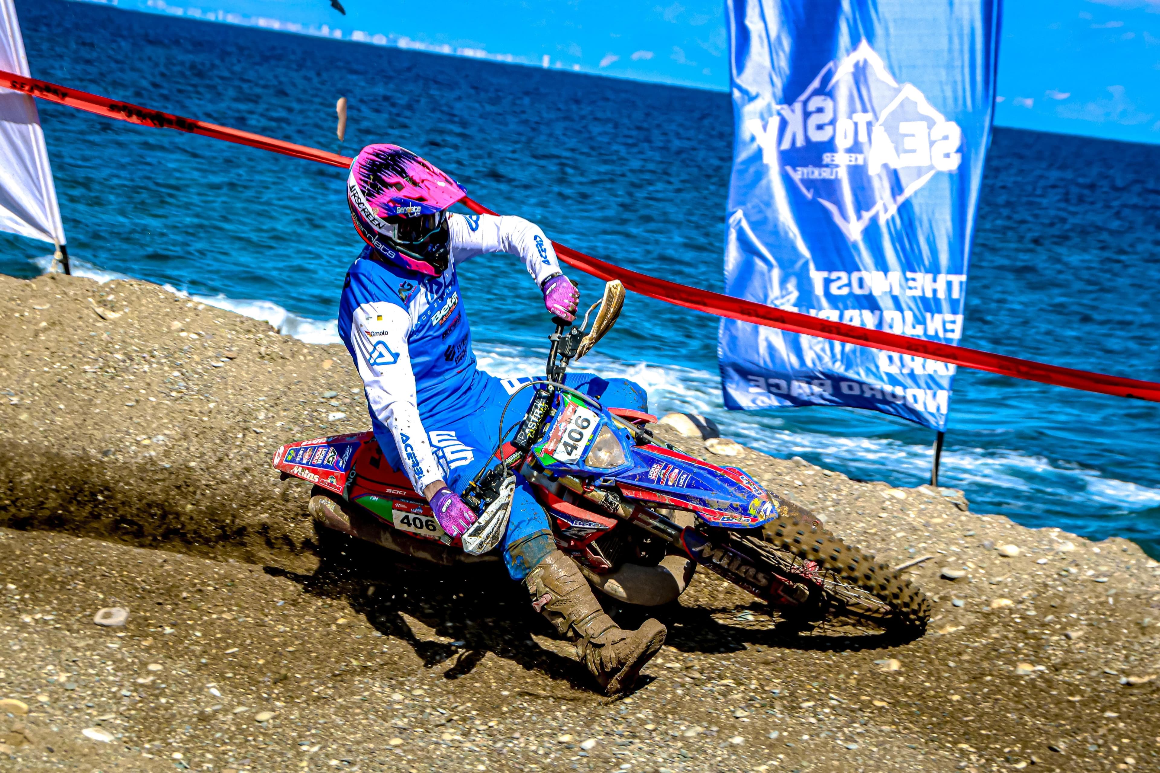 Motocross-Fahrer mit pinkem Helm steuert sein blaues Motorrad über eine sandige Piste am Meer.