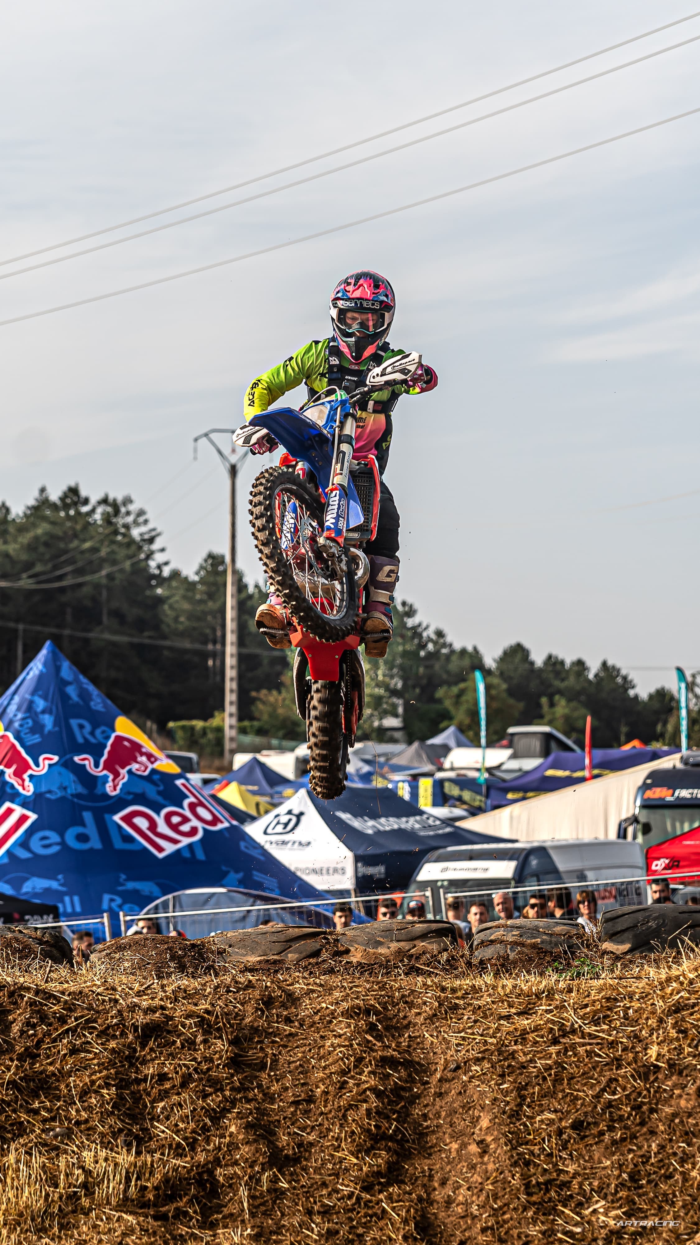 Ein Motocross-Fahrer in bunter Ausrüstung springt mit seinem blau-roten Motorrad hoch über einen Erdhügel.