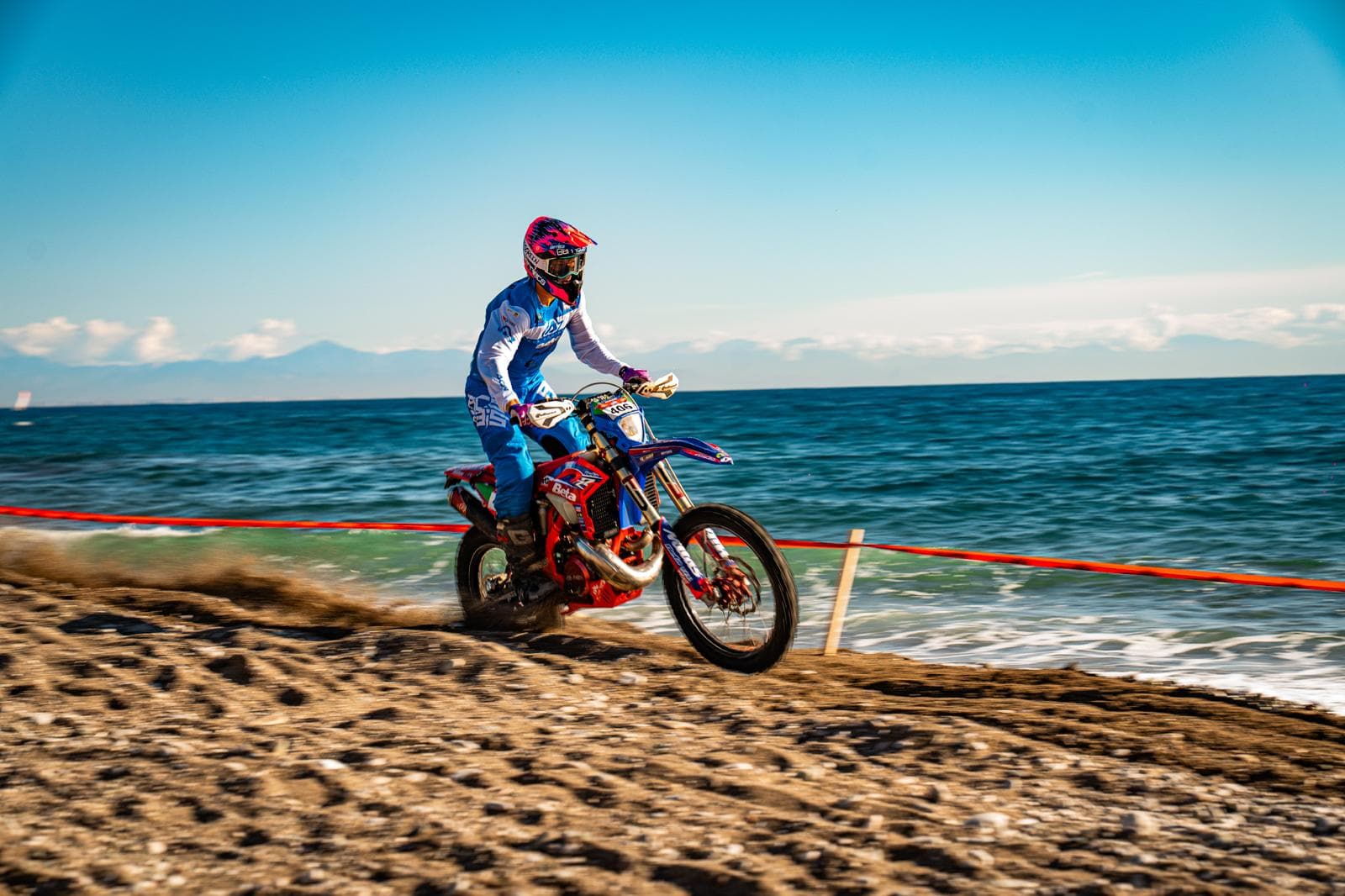 Motocross-Fahrer in blauer Kleidung rast über einen Kieselstrand am Ufer des strahlend blauen Meeres.