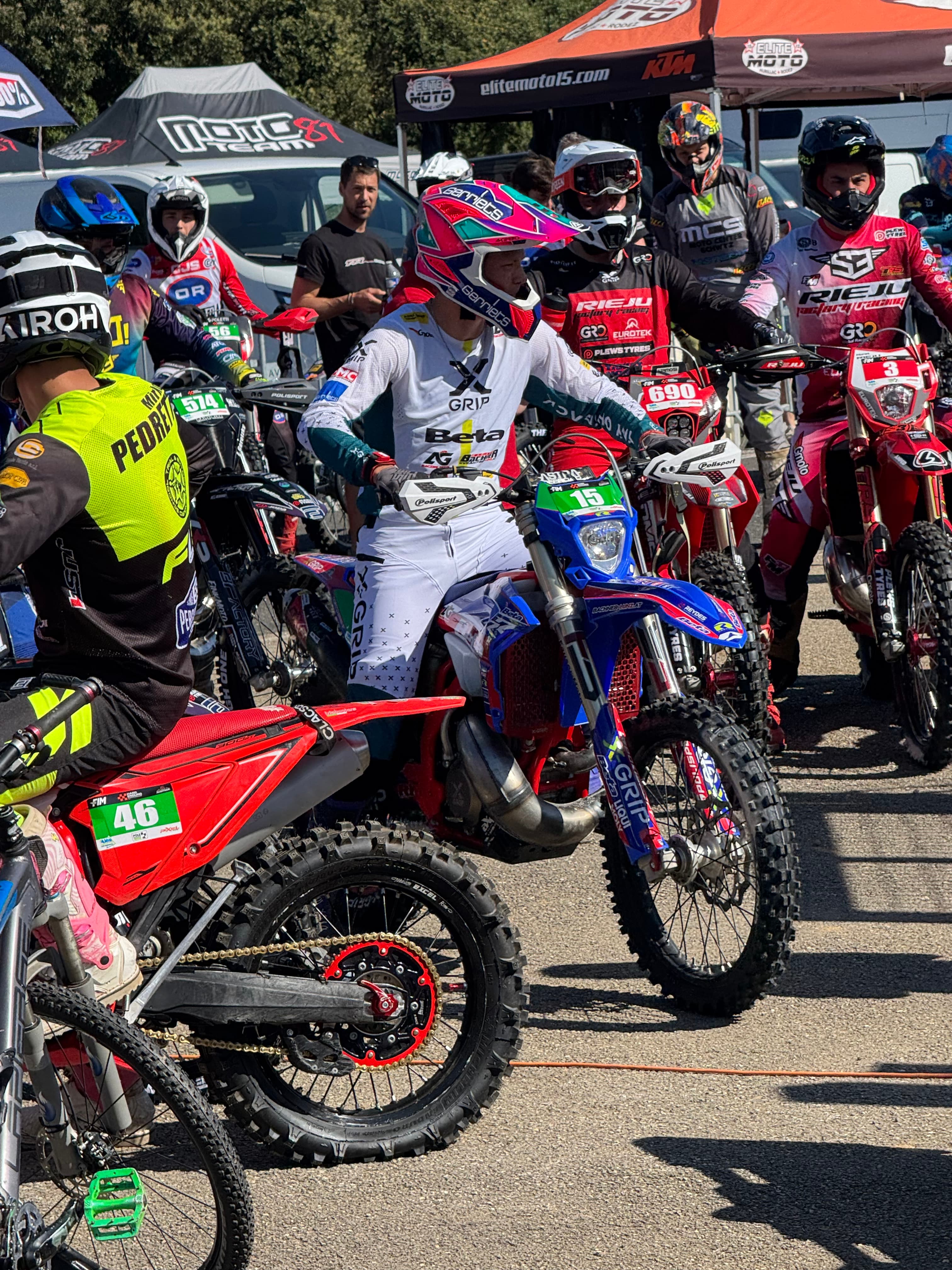 Mehrere Motocross-Rennfahrer in voller Montur posieren mit ihren farbenfrohen Motorrädern bei strahlendem Sonnenschein.