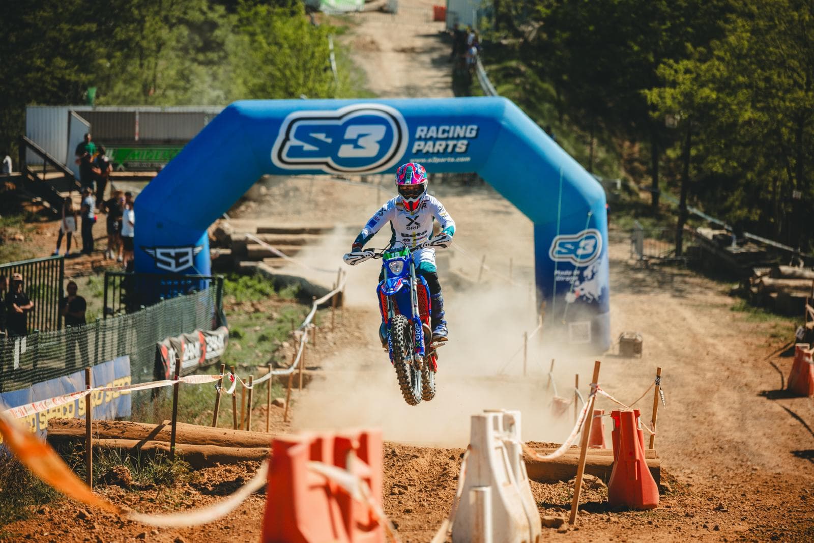Motocross-Fahrer springt über ein Hindernis auf einer staubigen Rennstrecke vor einem blauen Werbebogen.