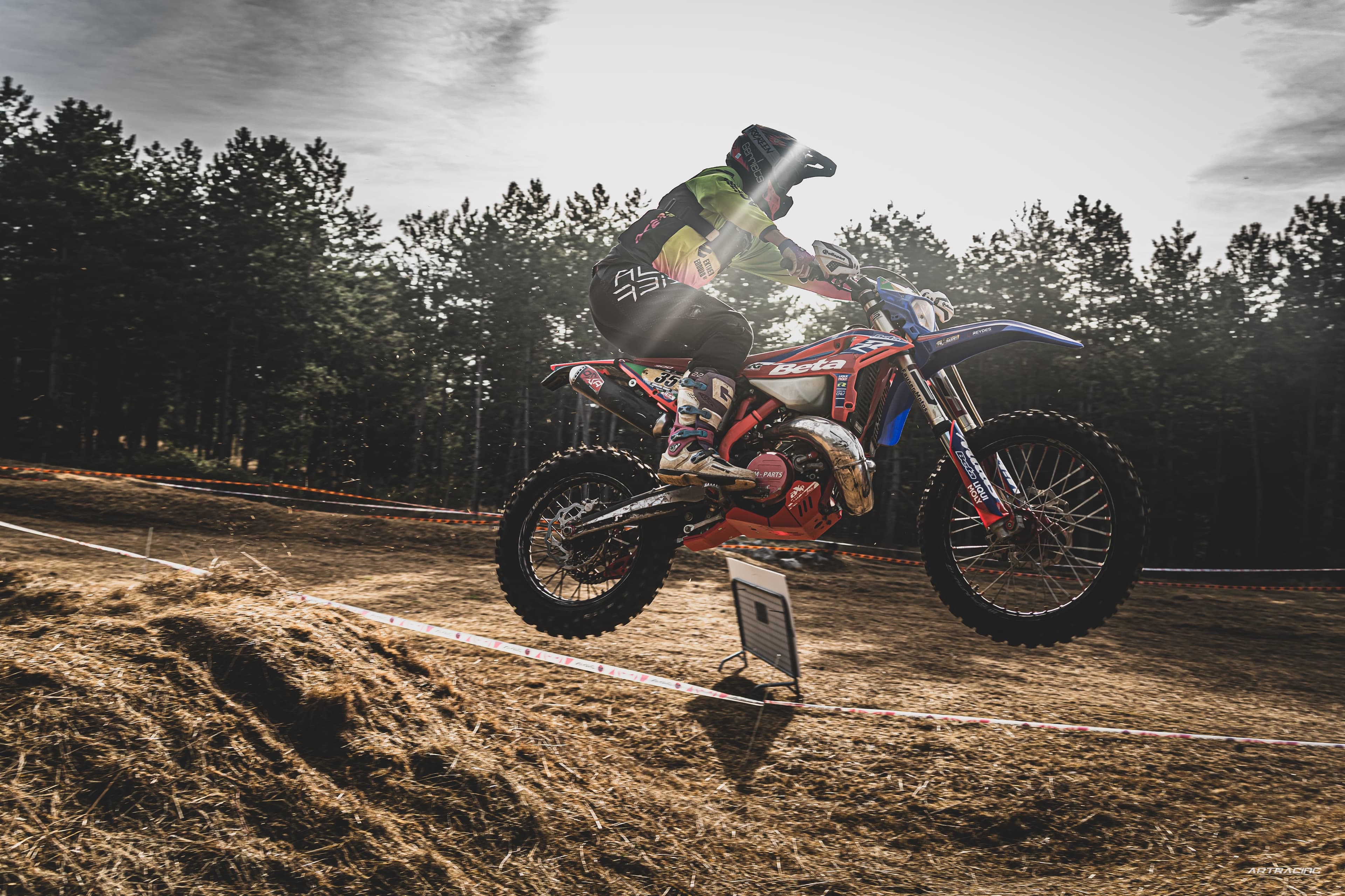 Motocross-Fahrer springt mit einer Beta-Maschine über einen Erdhügel vor einem dichten Nadelwald.
