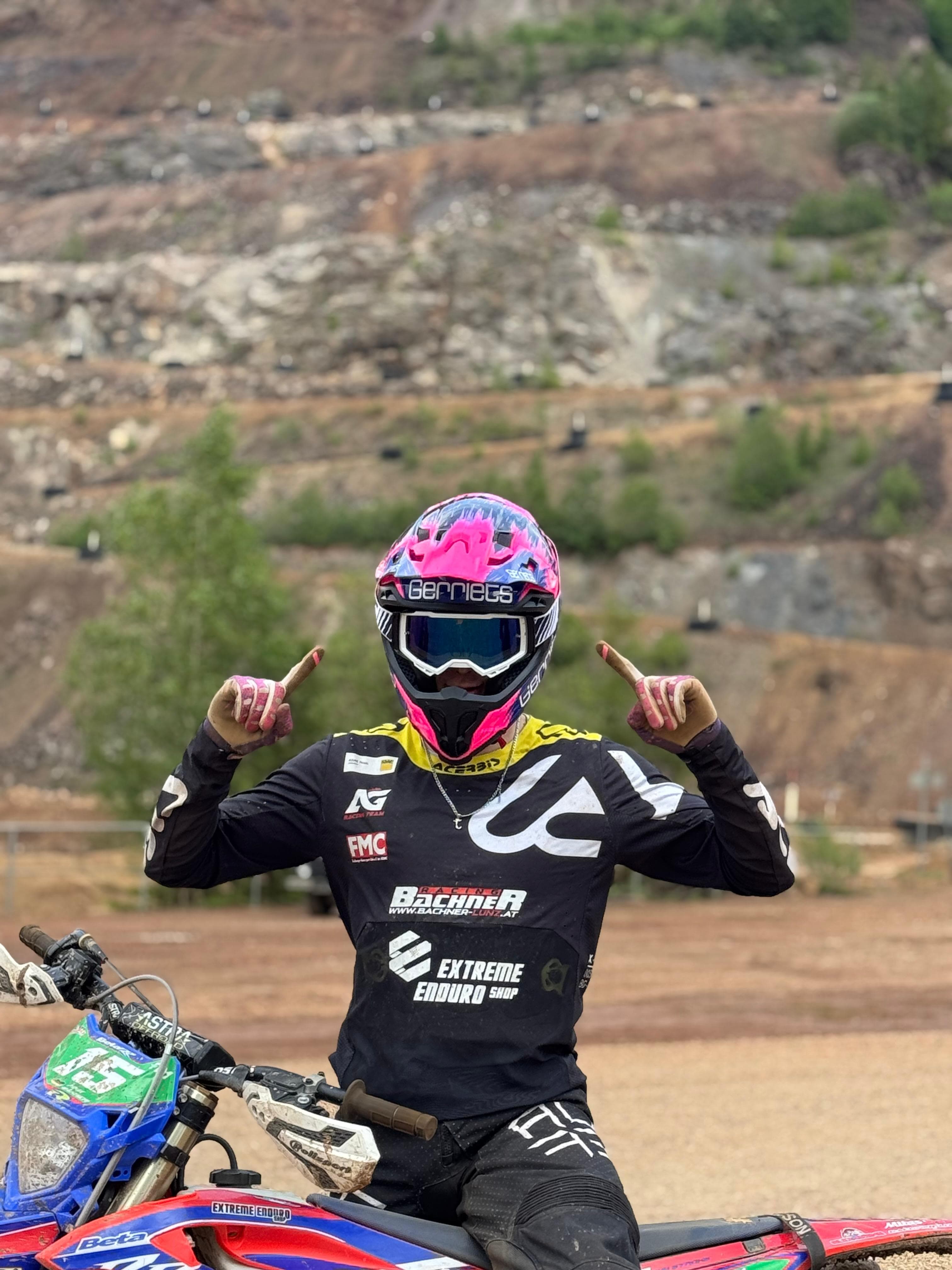 Motocross-Fahrer mit pinkem Helm zeigt triumphierend nach oben, sitzend auf seinem Motorrad vor Felskulisse.