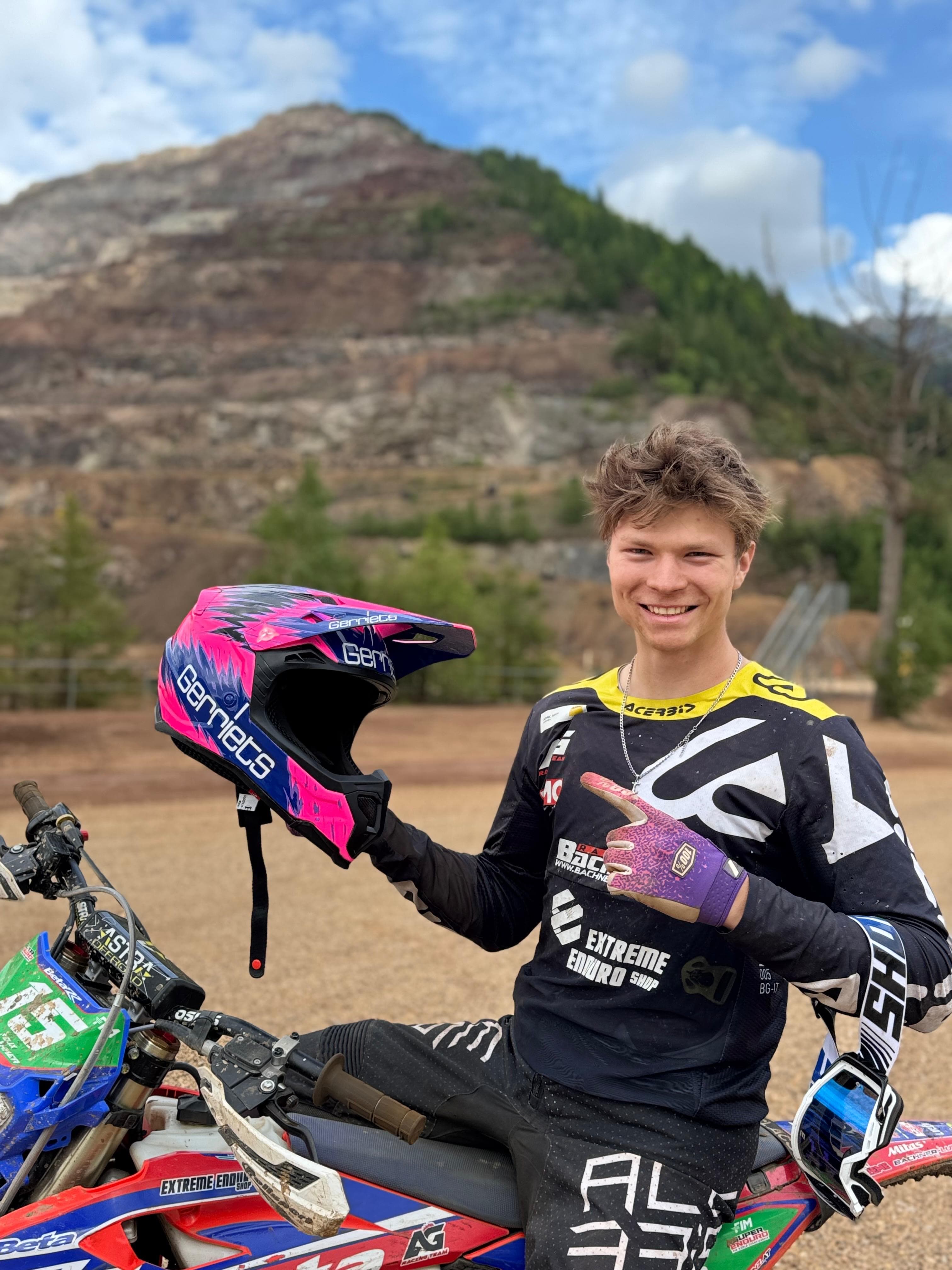 Lächelnder Motocross-Fahrer auf seinem Motorrad hält einen pink-blauen Helm vor einer felsigen Bergkulisse.